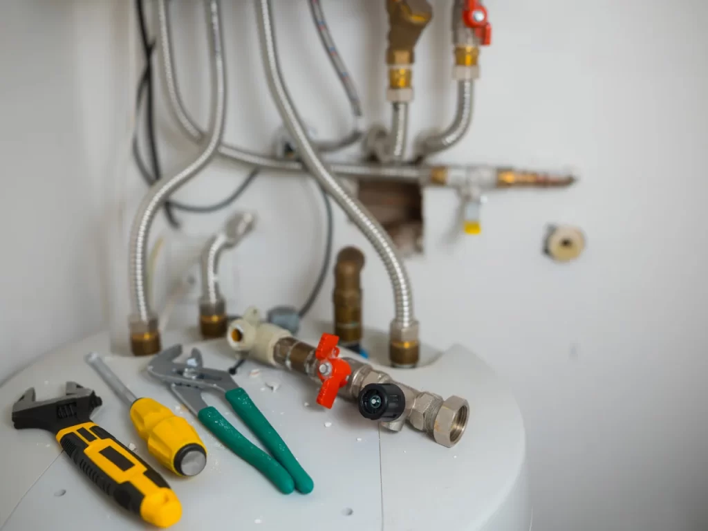A close-up of a water heater with various plumbing pipes above it and tools such as pliers, screwdrivers, and a wrench laid out on its surface.