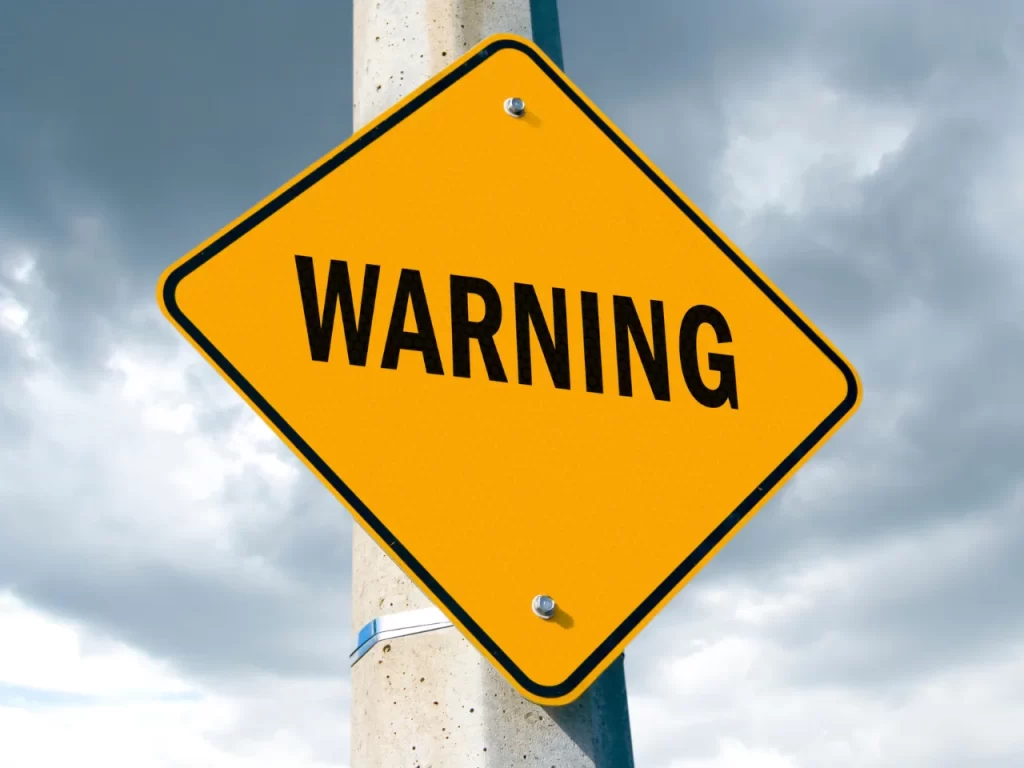A yellow diamond-shaped sign with the word WARNING in bold black letters, mounted on a pole against a cloudy sky background.