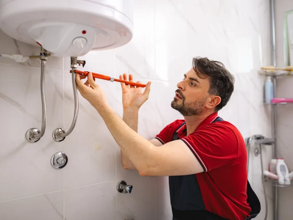 A man in a red shirt uses a wrench to fix pipes under a water heater mounted on a tiled bathroom wall.