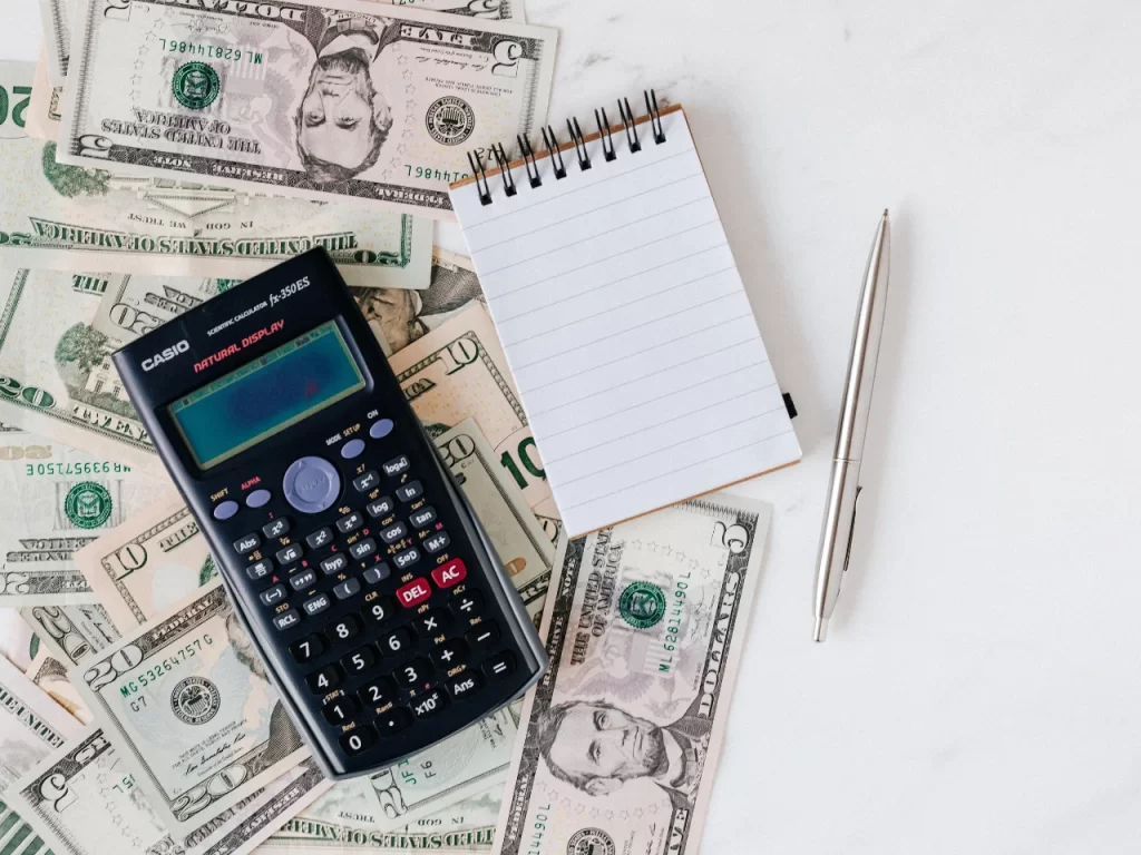 A scientific calculator, a blank notepad, and a silver pen are placed on top of scattered U.S. dollar bills on a white surface.