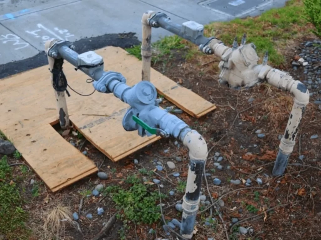 Two large, weathered metal and PVC pipes with valves and meters are installed outdoors on uneven ground, surrounded by gravel, grass, and two plywood boards.