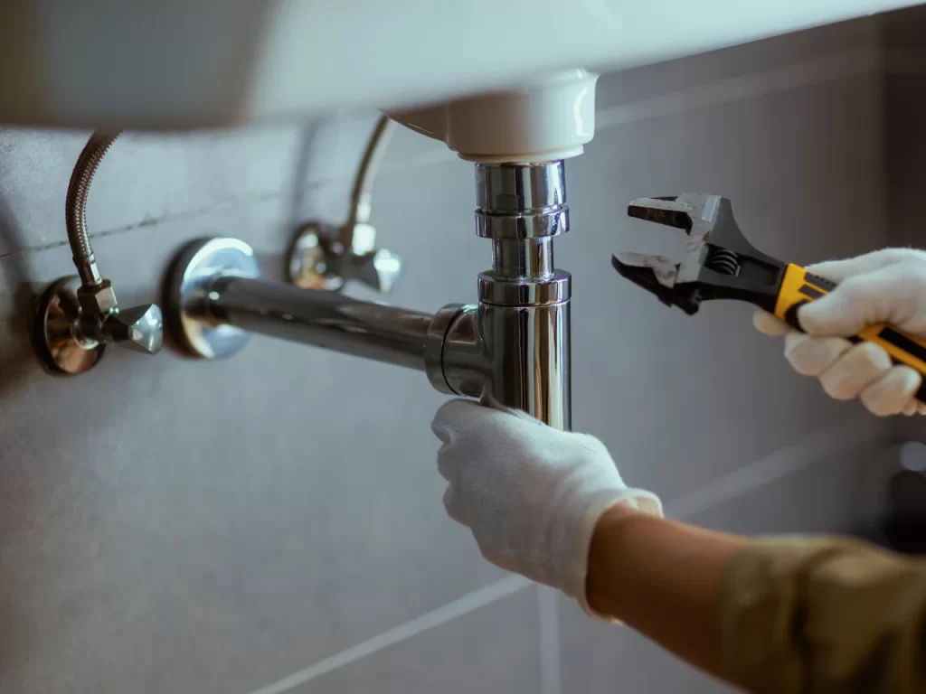 A person wearing white gloves uses a wrench to tighten or repair the metal pipe under a sink in a tiled bathroom.