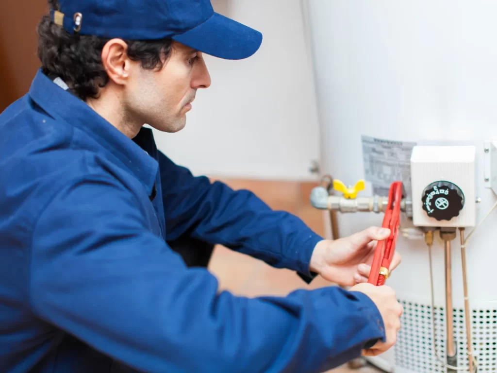 A worker in blue uniform and cap uses a red tool to adjust a valve on a white water heater, focusing intently on the task.