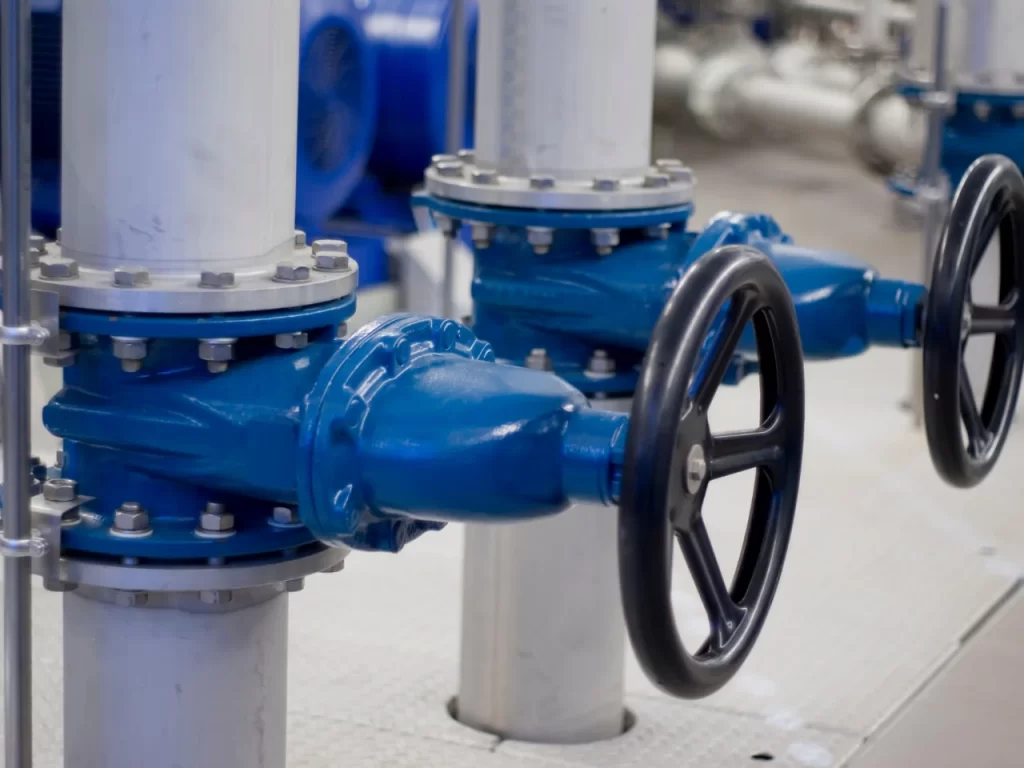 Close-up of two blue industrial pipe valves with black handwheels, attached to white vertical pipes in a clean indoor facility. The background shows additional pipes and equipment.