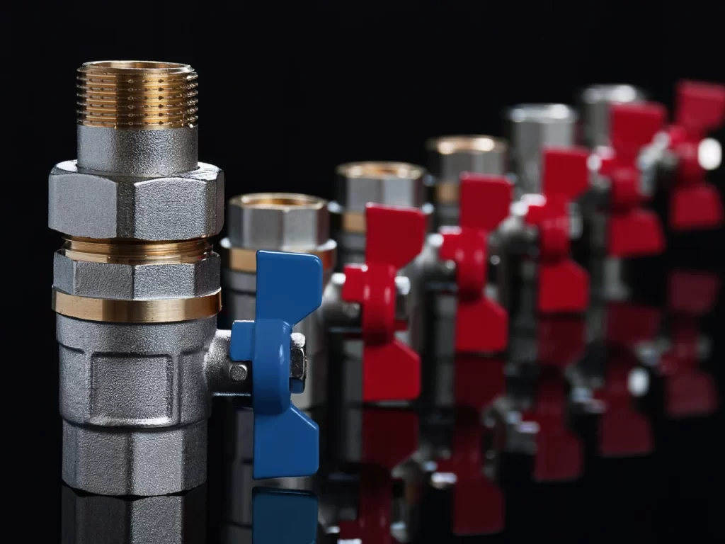 A row of metal plumbing valves with blue and red butterfly handles, arranged in a line on a reflective black surface. The closest valve has a blue handle; the others have red handles and fade into the background.