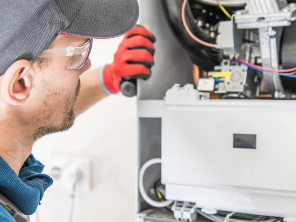 A technician wearing safety glasses, a cap, and red gloves inspects the internal components of a heating or HVAC unit, holding a tool and examining the machinery closely.