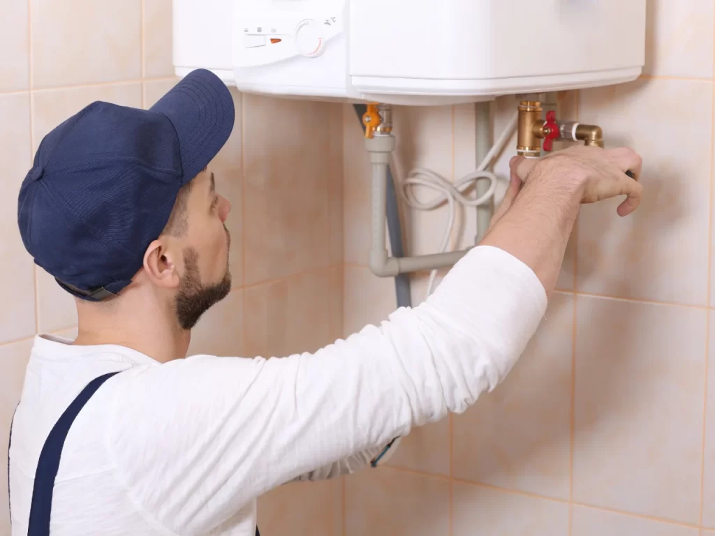 A plumber wearing a blue cap and white shirt adjusts pipes on a wall-mounted water heater in a tiled room.