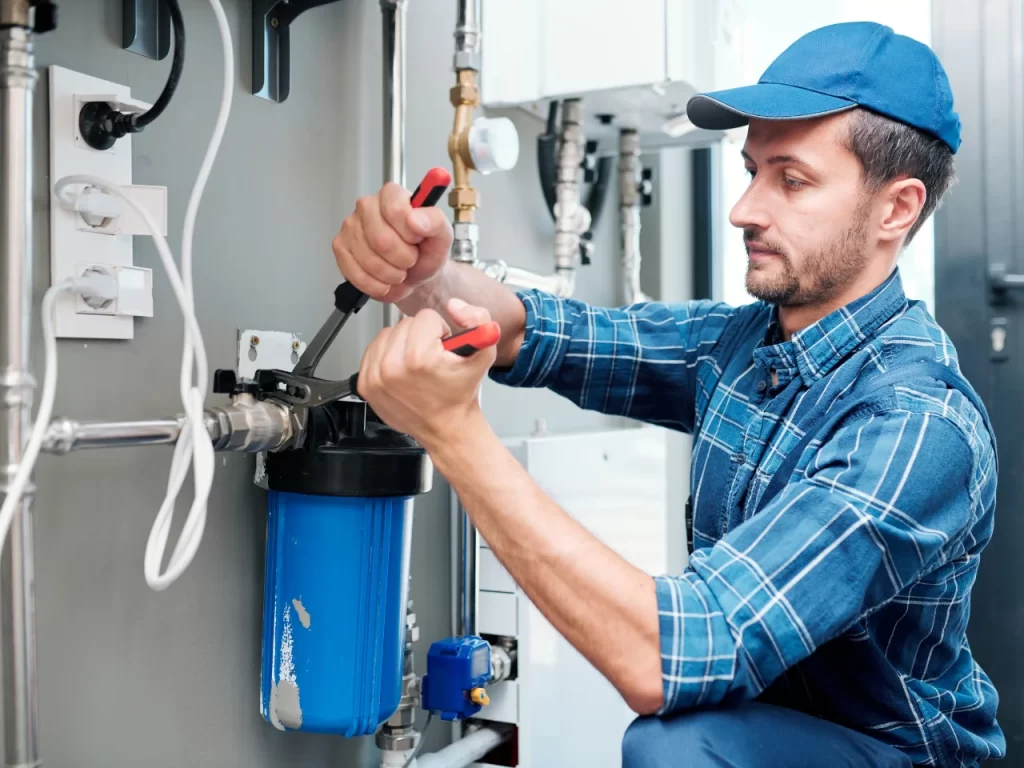 A plumber in a blue cap and plaid shirt uses tools to work on a blue water filtration system attached to a wall with various pipes and valves.