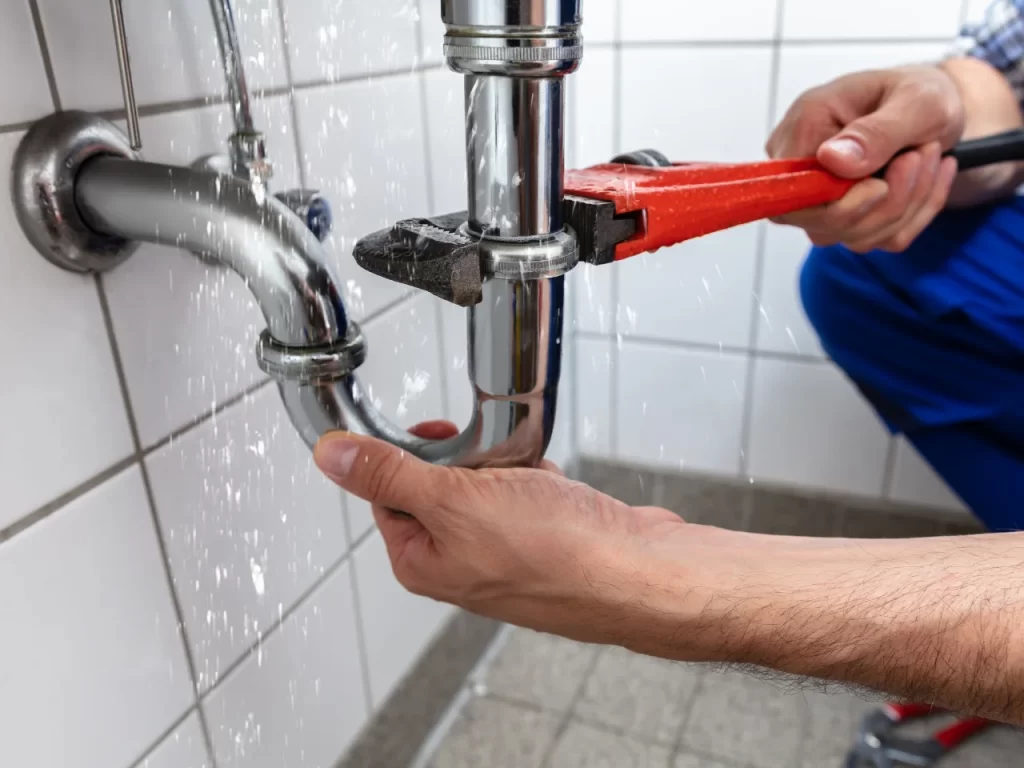 A person uses a red pipe wrench to fix a leaking chrome pipe under a sink, with water spraying out onto tiled walls and floor.