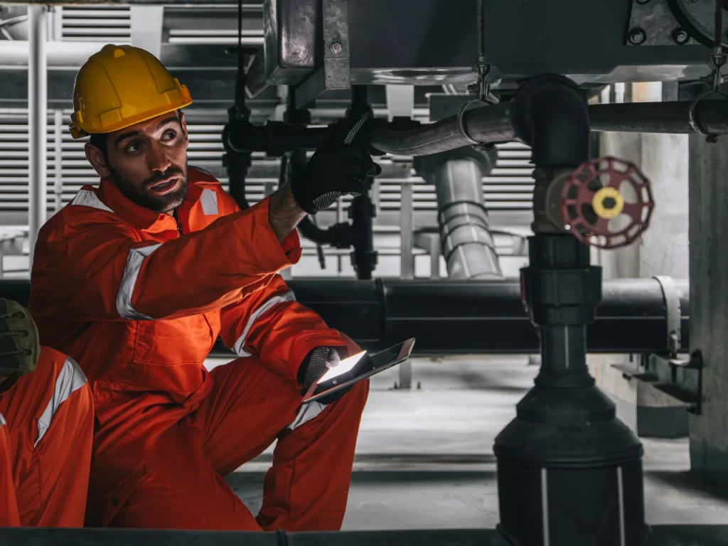 A worker in an orange jumpsuit and yellow hard hat crouches indoors, holding a tablet and pointing upwards at industrial pipes, appearing to inspect or discuss equipment.
