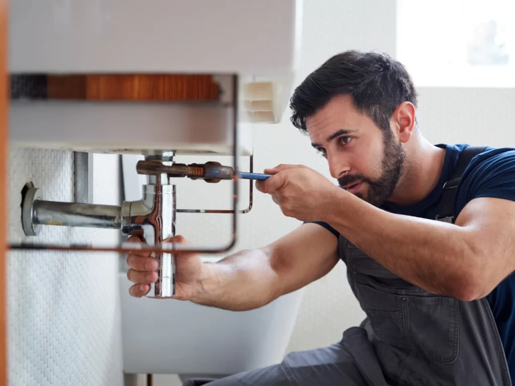 A plumber wearing overalls uses a wrench and screwdriver to fix pipes under a sink, focusing intently on his work.