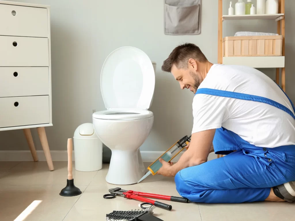 A man in blue overalls kneels next to a toilet, sealing its base with a caulking gun. Tools, including a plunger and wrenches, are on the floor nearby. The bathroom is tidy and well-lit.