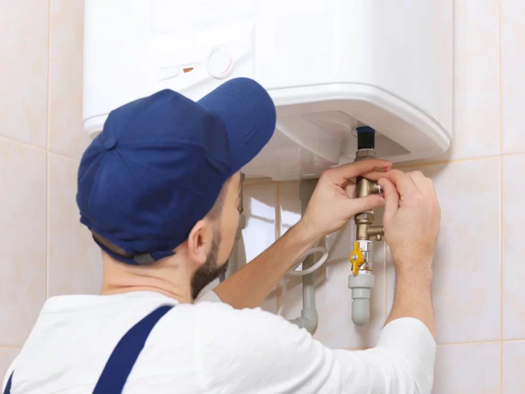 A person wearing a blue cap and uniform is repairing or adjusting valves on a water heater mounted on a tiled wall.