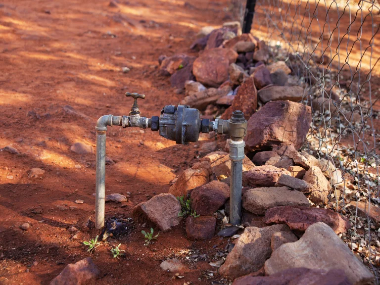 A water meter with a shut-off valve is installed on two metal pipes emerging from the ground, surrounded by reddish soil, rocks, and a wire fence—ideal for those needing drain repair or plumbing in Toronto.