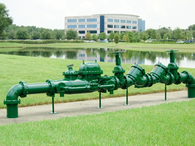 A large green water pipeline system with valves sits on a concrete pad in grassy landscaping, ideal for showcasing drain repair and plumbing in Toronto, with a pond and modern office building in the background.