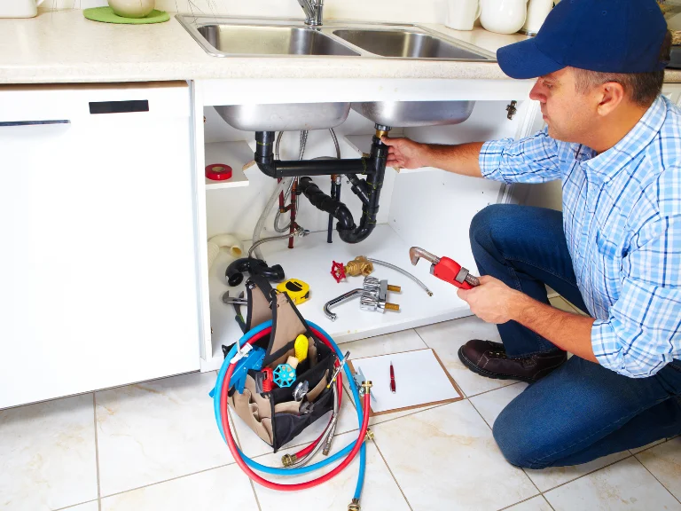 A plumber wearing a blue cap and plaid shirt kneels under a kitchen sink, handling drain repair and plumbing in Toronto. An open tool bag with various plumbing equipment sits nearby on the floor.