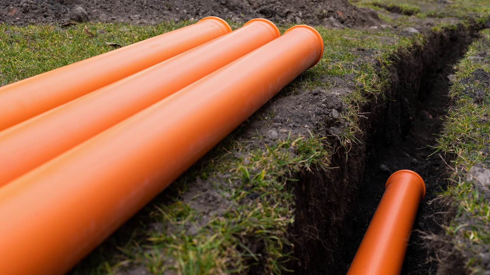 Four large orange plastic pipes are laid on grass next to a trench, with one pipe partially placed inside the trench in the soil, suggesting installation or construction work.