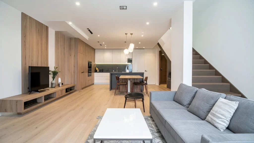 Modern open-plan living space with light wood floors, a gray sofa, white coffee table, TV on a wooden stand, dining area, and kitchen in the background. A staircase leads to an upper floor on the right.