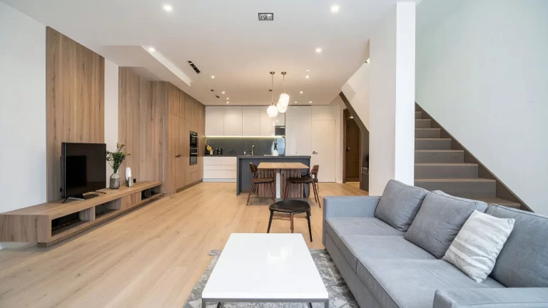 Modern open-plan living space with light wood floors, a gray sofa, white coffee table, TV on a wooden stand, dining area, and kitchen in the background. A staircase leads to an upper floor on the right.