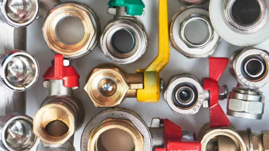 Close-up view of various metal plumbing fittings and valves, some with red, yellow, and green handles, arranged in a tightly packed pattern.