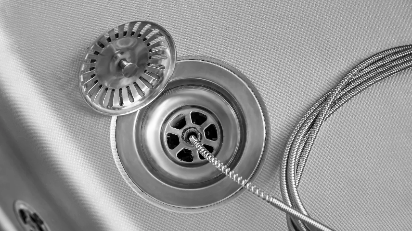 Close-up of a stainless steel sink drain with a strainer partially covering it and a flexible metal hose placed nearby. The image is in black and white.