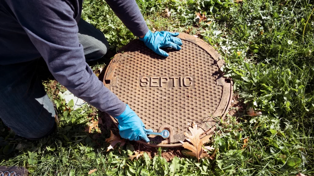 A person wearing blue gloves uses a wrench to open a round septic tank cover labeled SEPTIC, surrounded by grass and fallen leaves.