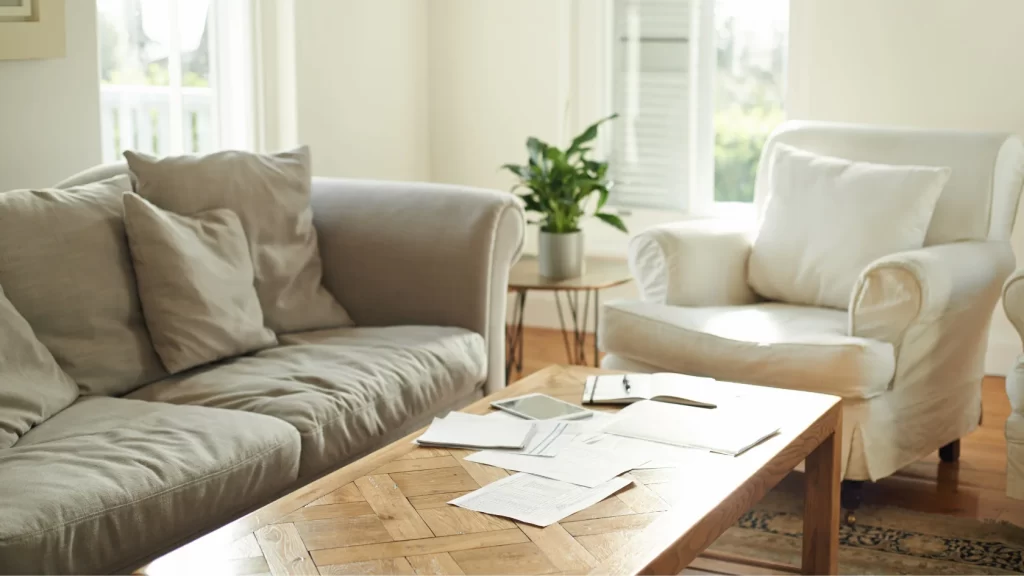 A sunlit living room with a beige sofa, a white armchair, a wooden coffee table with papers, a notebook, a phone, and a pen, and a potted plant near a window with white shutters.