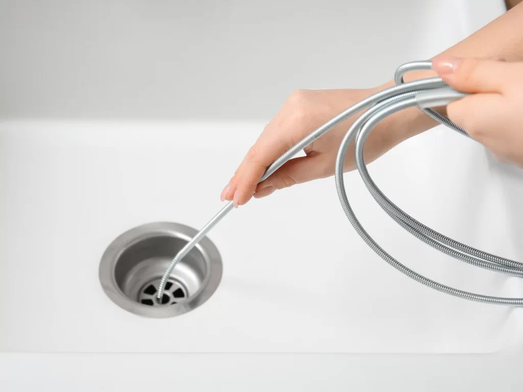 A person’s hands are inserting a flexible metal drain snake into a kitchen sink drain, likely to unclog or clean the pipe.