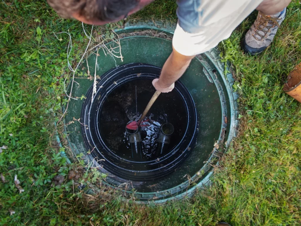 A person wearing outdoor shoes and gloves uses a tool to inspect or maintain equipment inside an open green septic tank or utility access hatch on a grassy lawn.