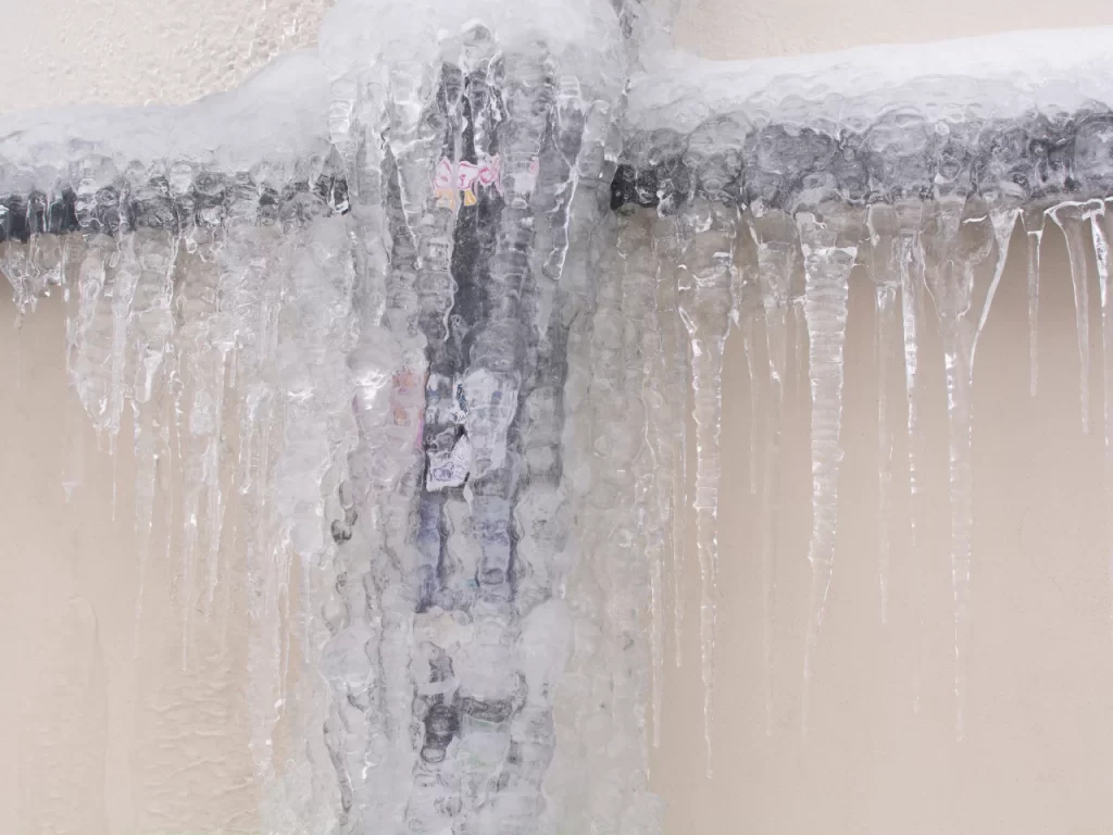 A metal water pipe covered in thick layers of ice and icicles is attached to a light-colored wall, showing signs of freezing weather.