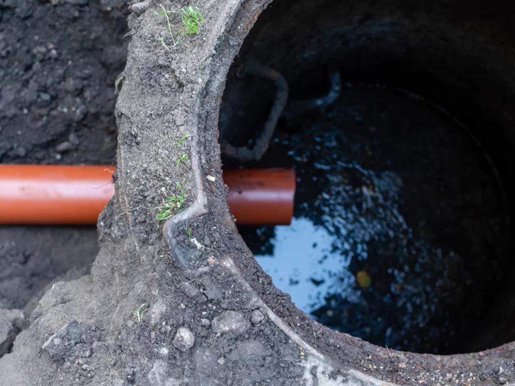 A close-up of an open, partially buried concrete manhole with a brown pipe leading into it. The surrounding soil is visible, and there is water inside the manhole reflecting the sky.