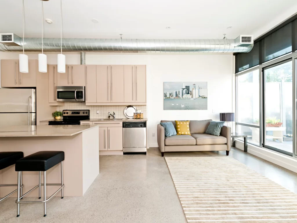Modern open-concept kitchen and living room with beige cabinets, stainless steel appliances, a gray sofa with yellow and blue pillows, large windows, a striped rug, and industrial exposed ductwork.