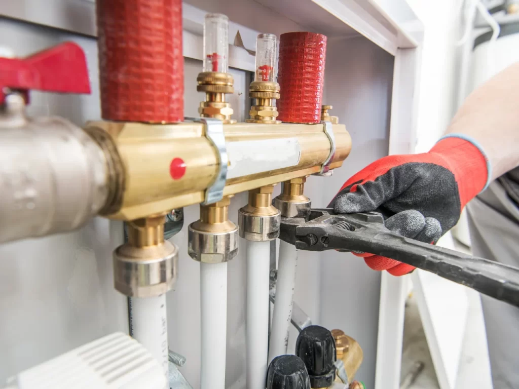 A person wearing gloves uses a wrench to tighten a fitting on a brass manifold with pipes and valves, likely part of a heating or plumbing system.