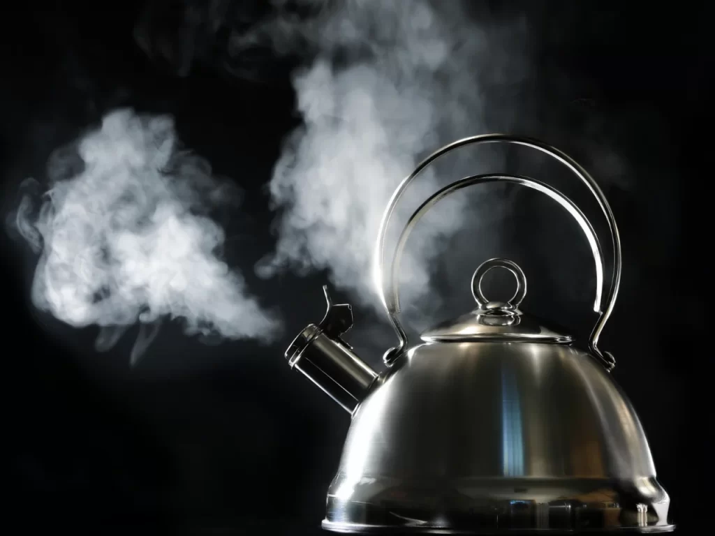 A stainless steel kettle with steam billowing from its spout against a dark background. The steam is visible, creating a contrast with the shiny metal surface of the kettle.