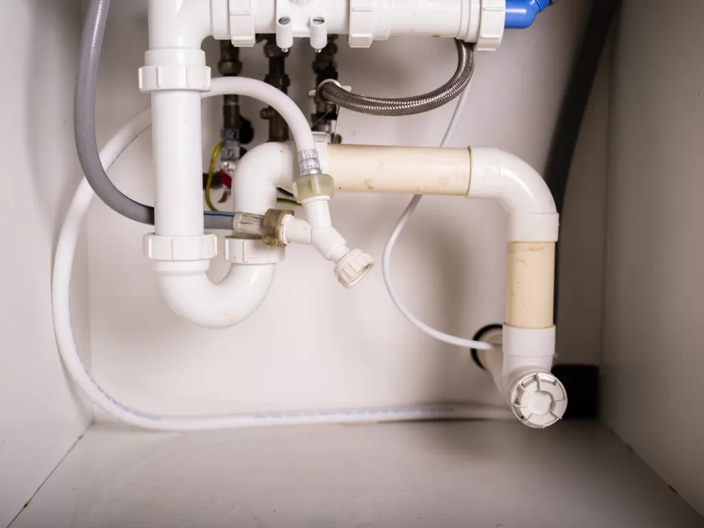 White PVC plumbing pipes, connectors, and hoses under a sink, showing a P-trap and various fittings against a white cabinet background.
