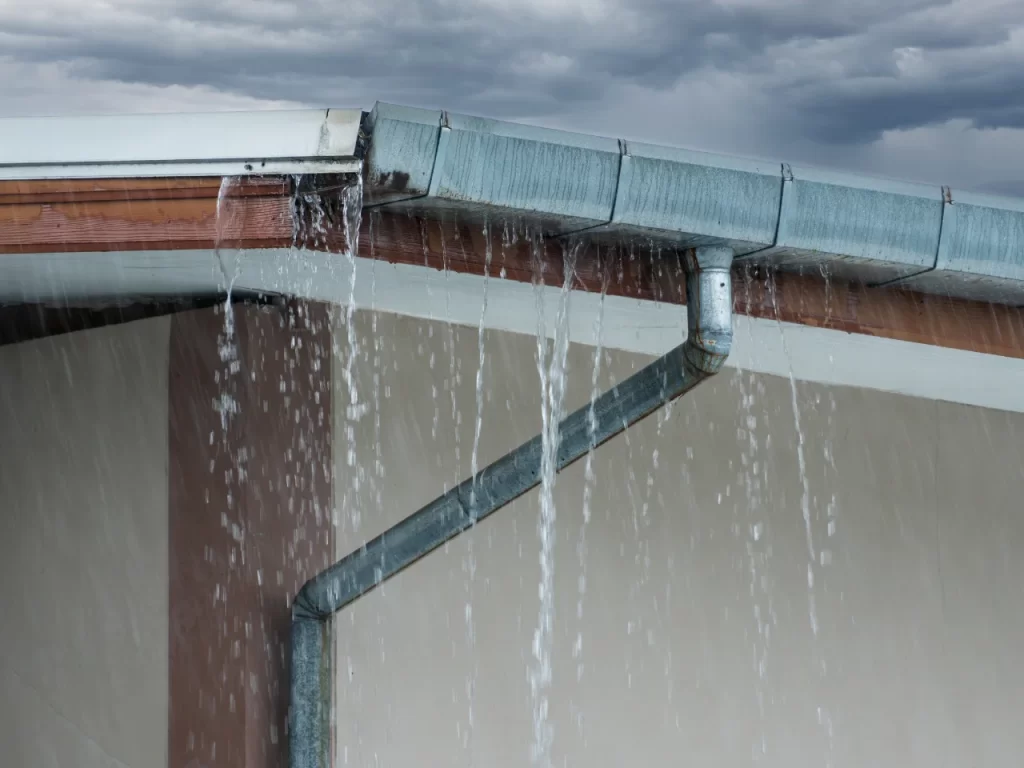 A metal gutter overflowing with rainwater spills over a roof’s edge against a cloudy sky, suggesting blockage—much like issues revealed in a faulty weeping tile diagram indicating poor drainage.