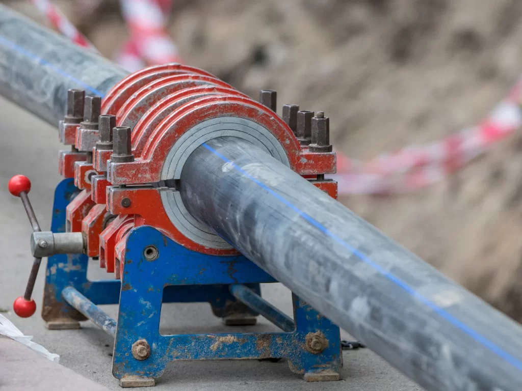 A close-up of a pipe fusion welding machine joining two large black plastic pipes at a construction site, with bolts and handles securing the pipes in place.