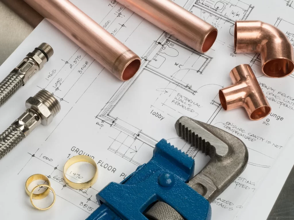 Copper pipes, fittings, a pipe wrench, and flexible connectors are arranged on top of an architectural floor plan, representing plumbing work or planning for a building project.