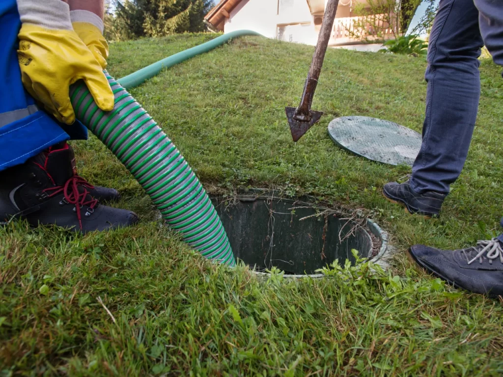 Two people in work boots are cleaning a septic tank; one holds a green hose inserted into the tank, while the other stands nearby with a shovel on a grassy lawn.