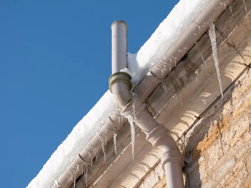 A snow-covered roof edge with a gutter and downspout, both coated in ice and icicles, against a clear blue sky. The building’s brick wall is partially visible below the roofline.
