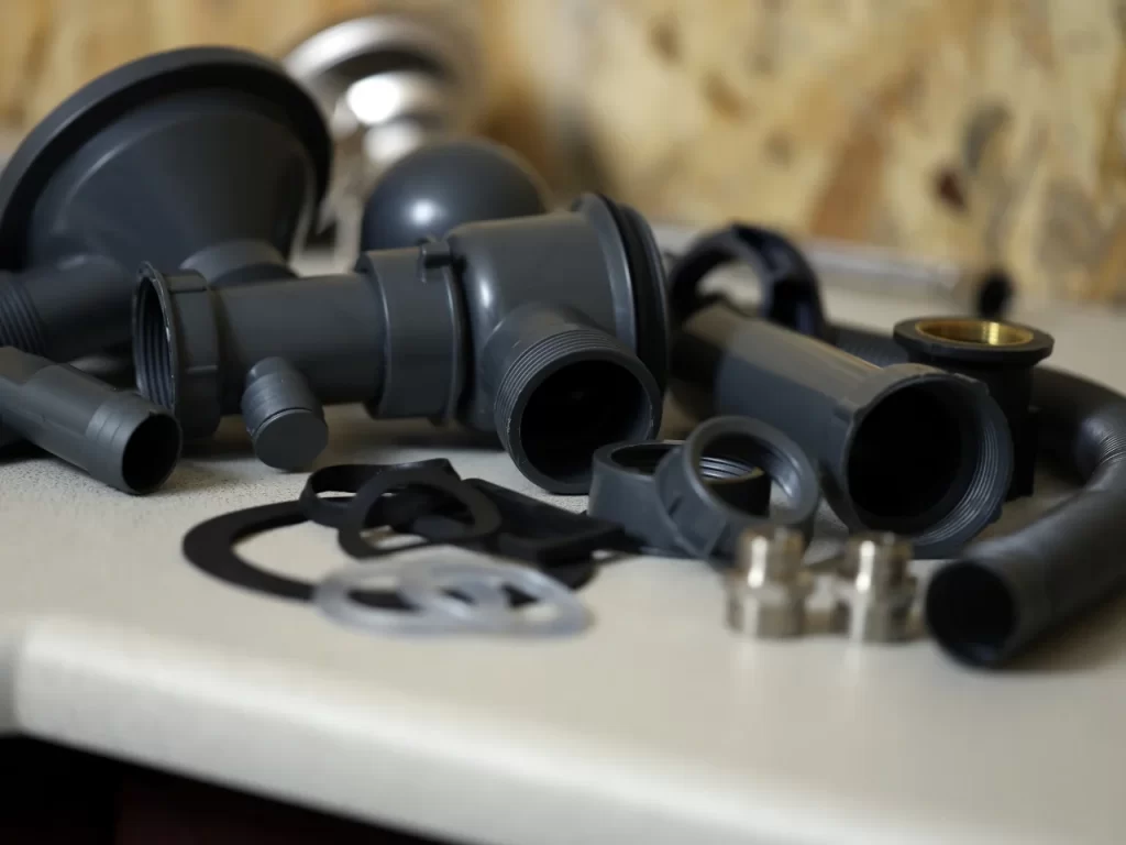 Assorted grey plastic plumbing pipe fittings, washers, and metal connectors are scattered on a white surface with a blurred, beige background.