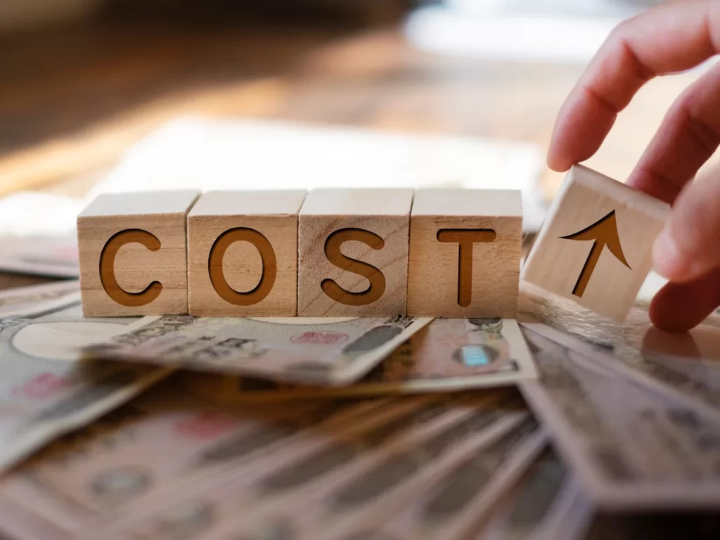 Wooden blocks spelling COST sit on top of scattered currency notes, with a hand placing a block showing an upward arrow, symbolizing rising costs or increasing expenses.