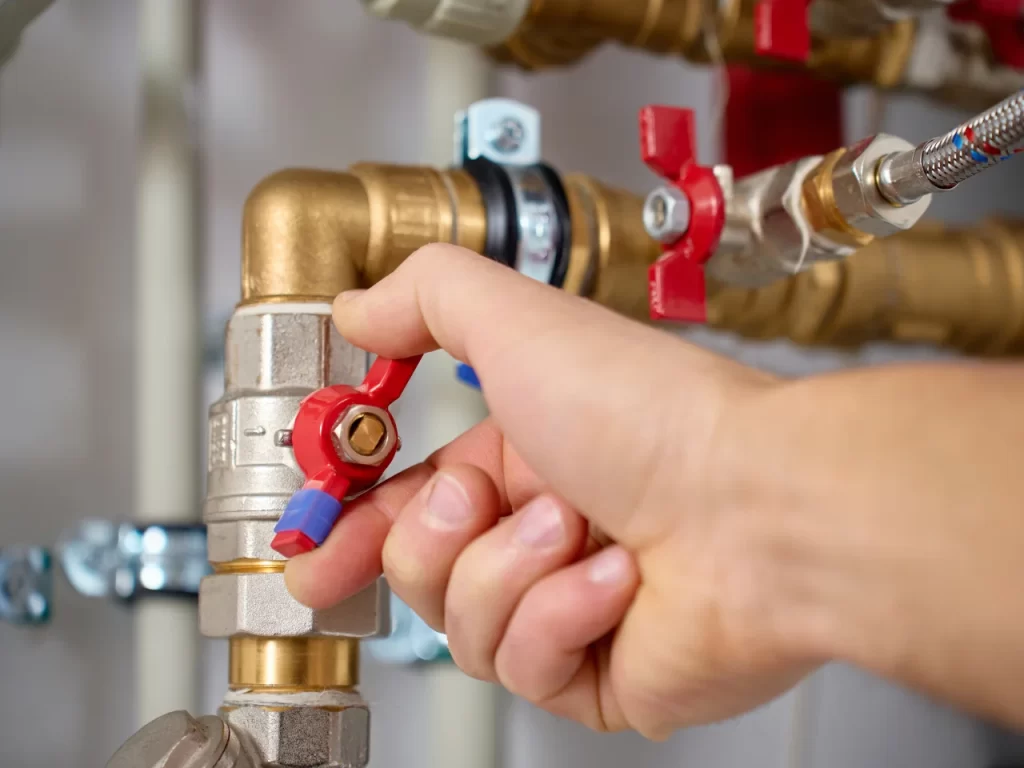 A close-up of a hand turning a red valve on a metal pipe, part of a plumbing system with several connected pipes and fittings visible in the background.