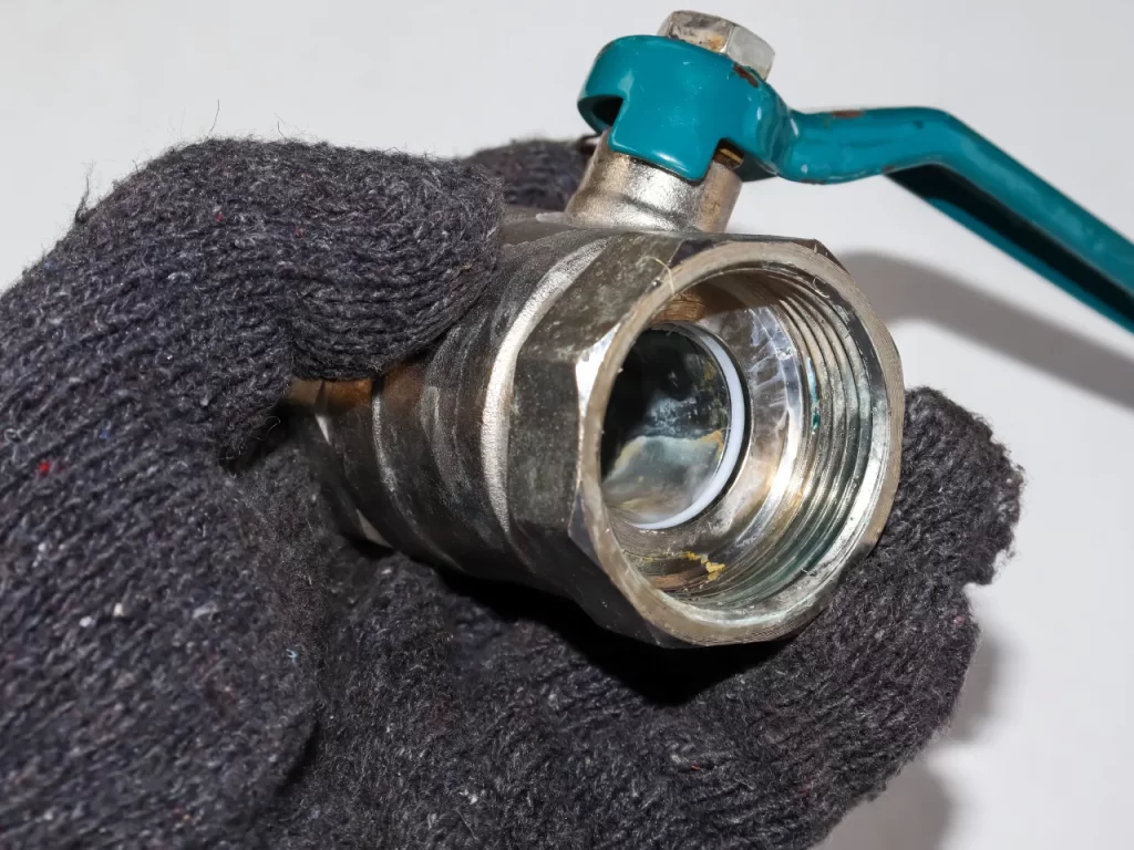 A gloved hand holding a metal ball valve with a teal handle, showing the open threaded end and internal ball mechanism. The background is plain and unfocused.
