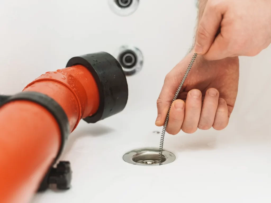 A person’s hand uses a plumbing snake to unclog a bathtub drain, with a red pipe and silver fixtures visible nearby.