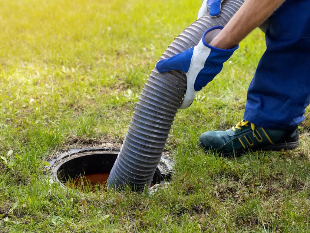A person wearing gloves and work shoes holds a large hose, inserting it into an outdoor septic tank opening surrounded by grass.