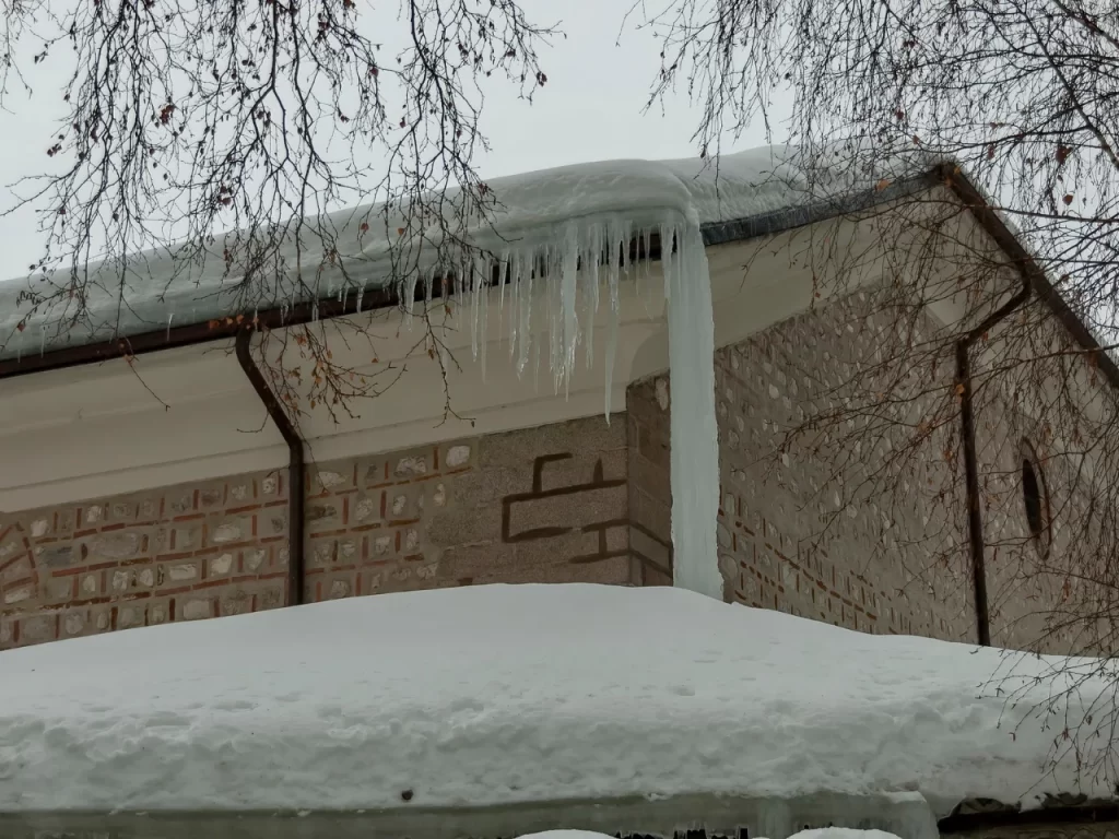 Large icicles hang from the edge of a snow-covered roof on a brick building. Snow piles up on the ground and bare tree branches frame the scene against a gray winter sky.