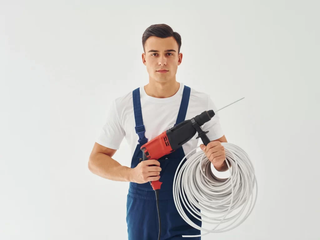 A man in blue overalls and a white t-shirt holds a red and black power drill in one hand and a large coil of white cable in the other, standing against a plain white background.