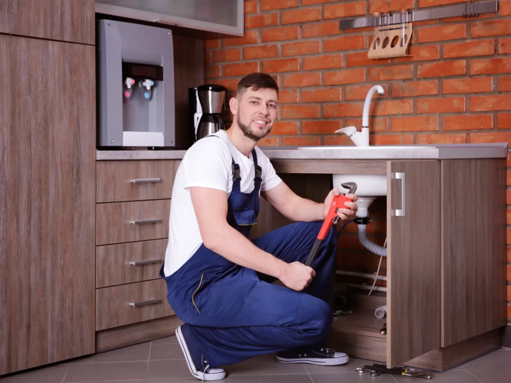 A plumber in blue overalls kneels on the kitchen floor, holding a red pipe wrench while working under a sink. He is smiling and surrounded by wooden cabinets and a brick wall.