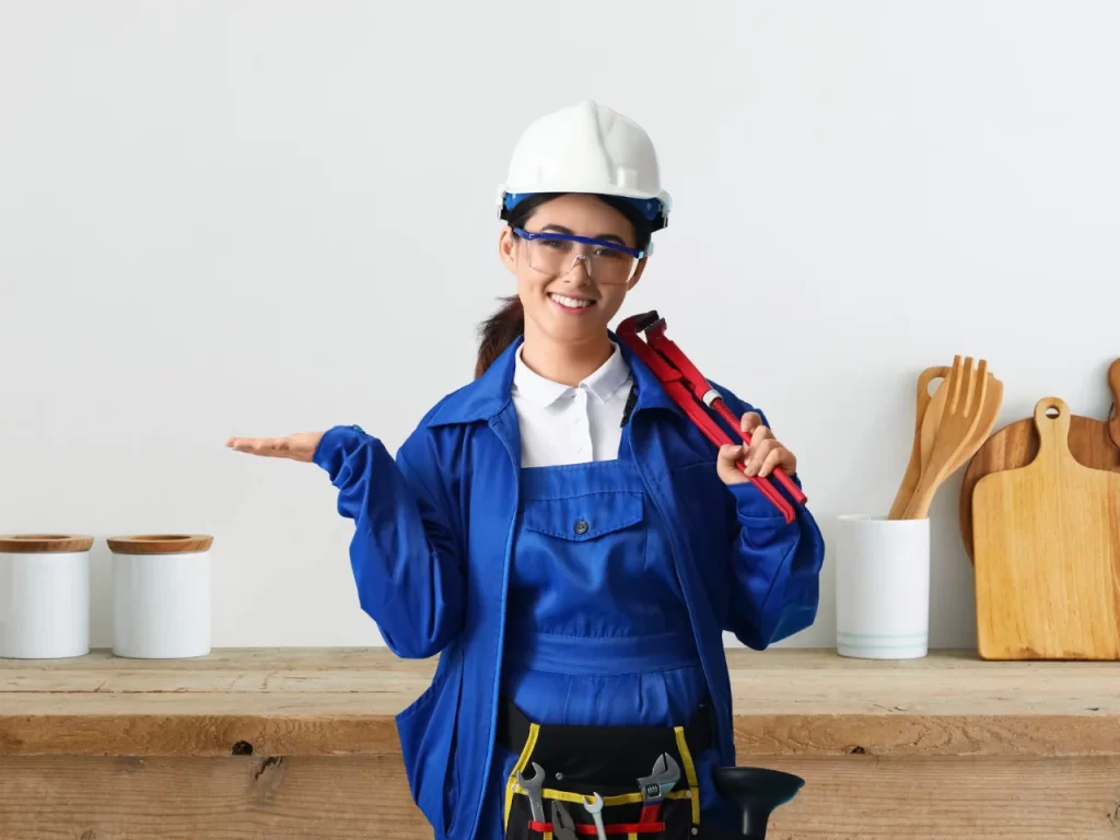 A smiling worker in blue coveralls, safety glasses, and a white hard hat holds a red pipe wrench over one shoulder and gestures with the other hand in a kitchen setting with utensils on a wooden counter behind her.